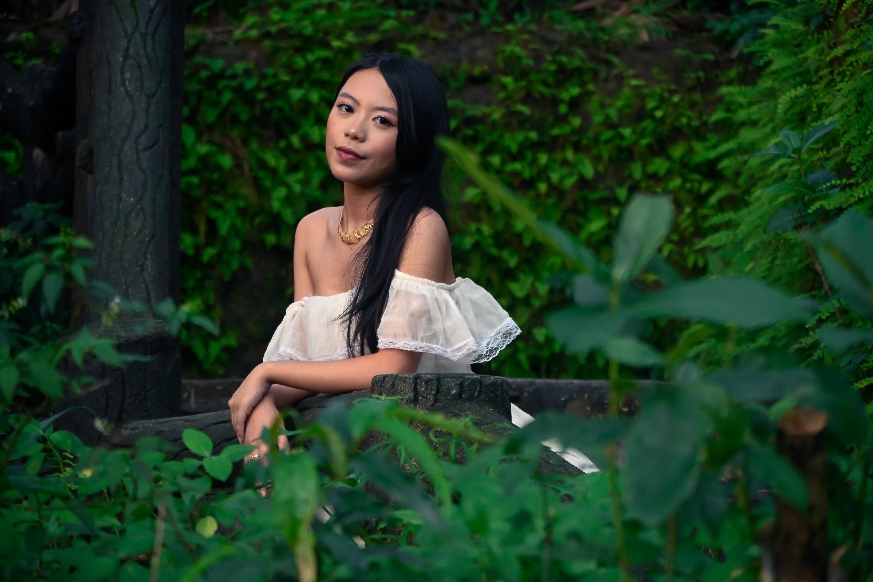 Portrait in an off-shoulder white gown beside a wooden garden rail, framed by green leaves.
