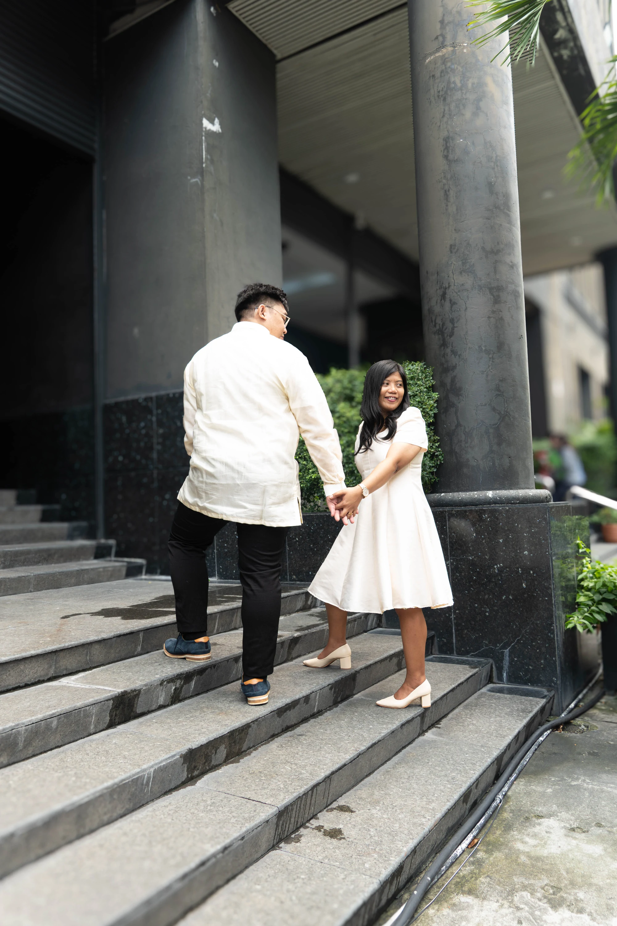 The couple holds hands on steps beside tall concrete columns.