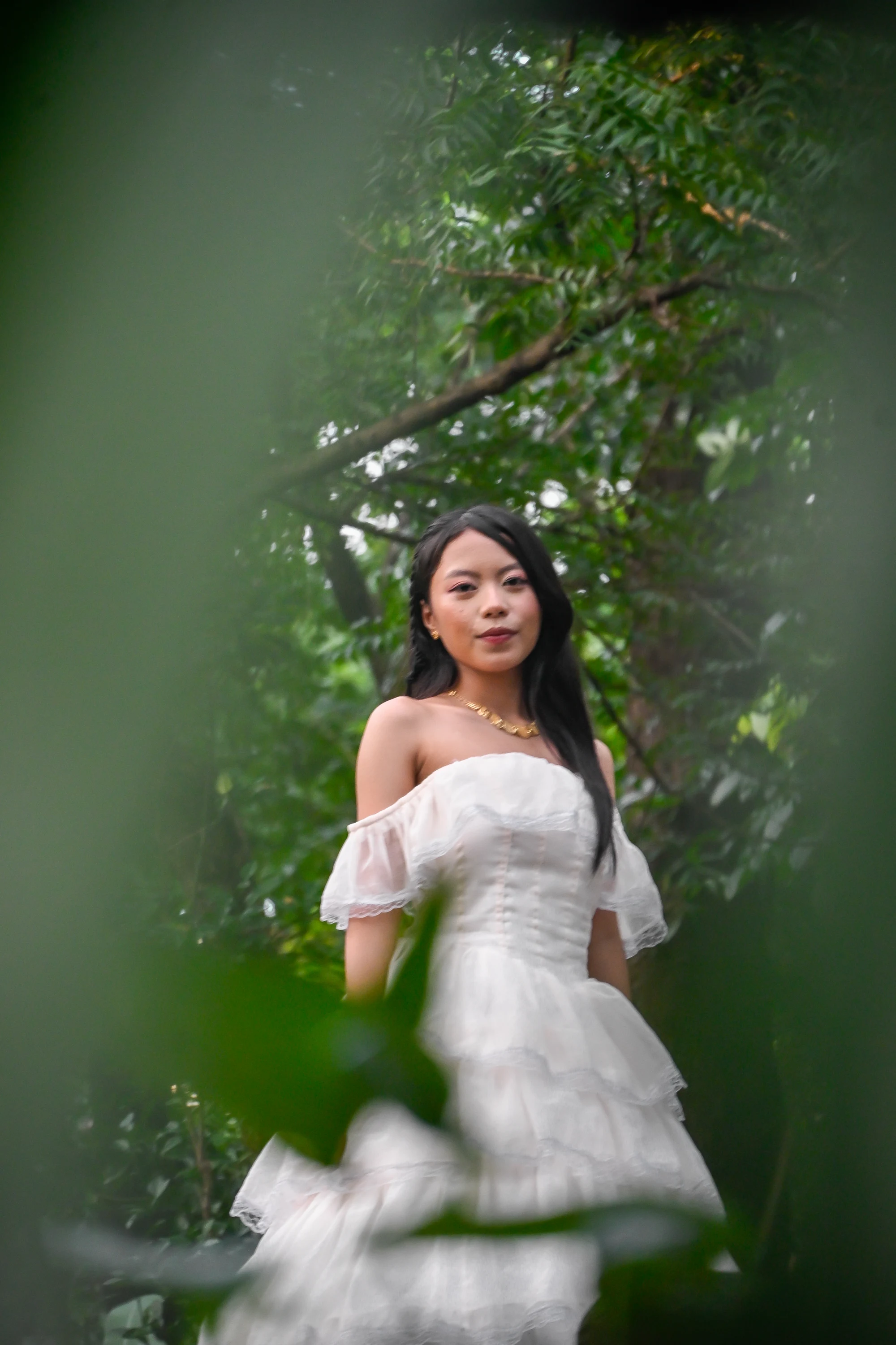 Standing portrait in a white gown framed by softly blurred foreground leaves.