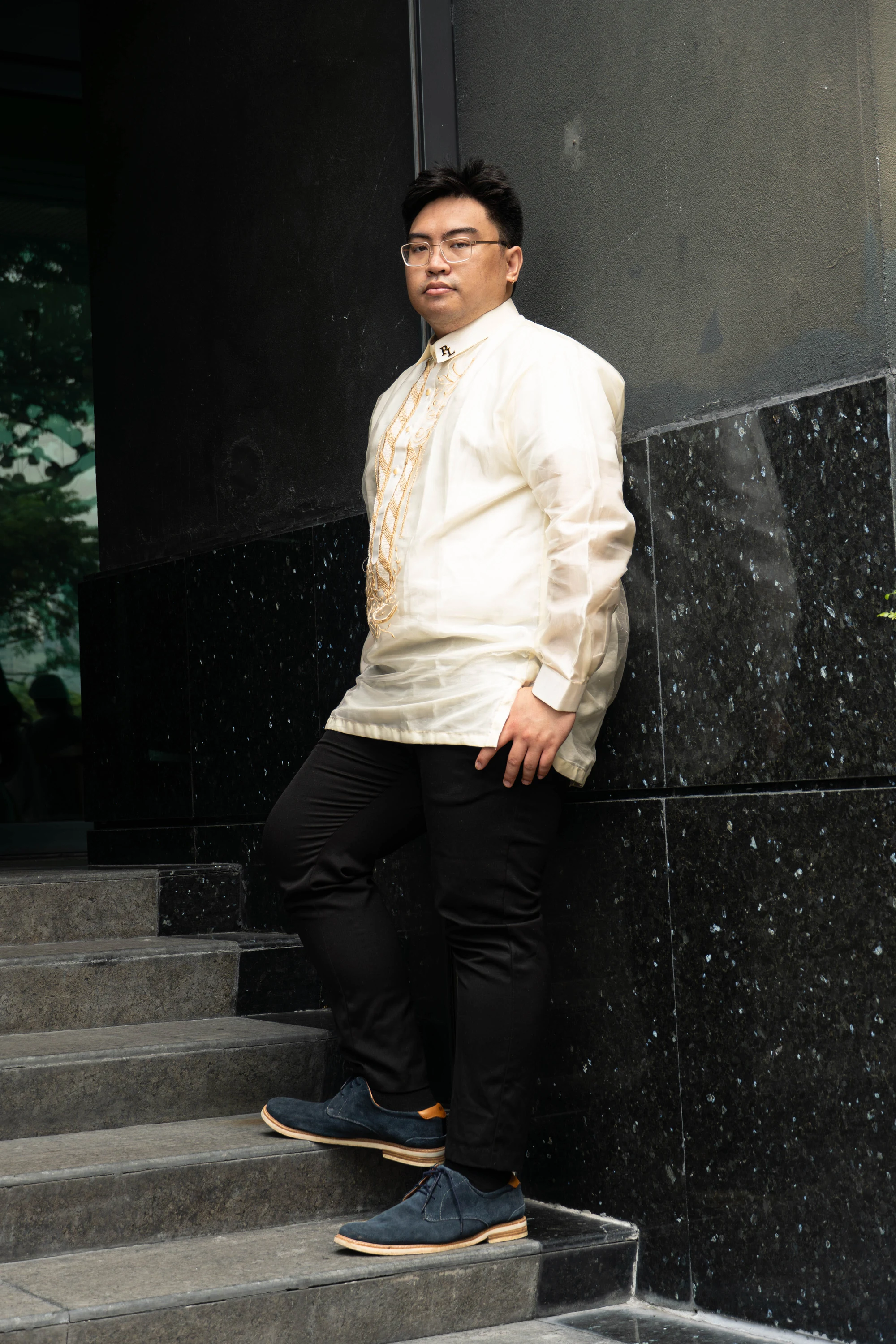 Portrait of the groom wearing an embroidered ivory barong beside a dark stone wall.