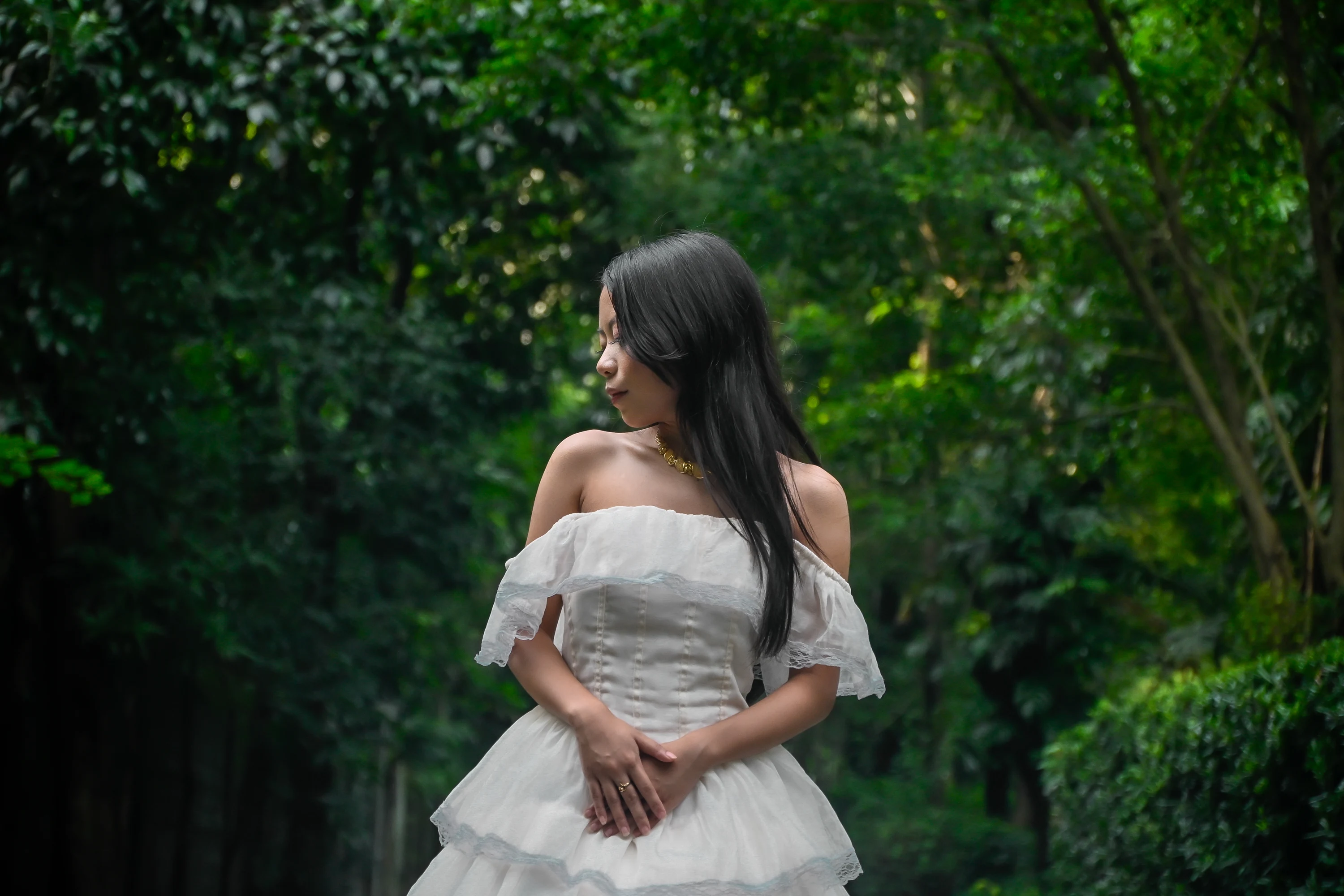 Portrait in a white gown with hands folded at her waist, looking to the side among leafy trees.