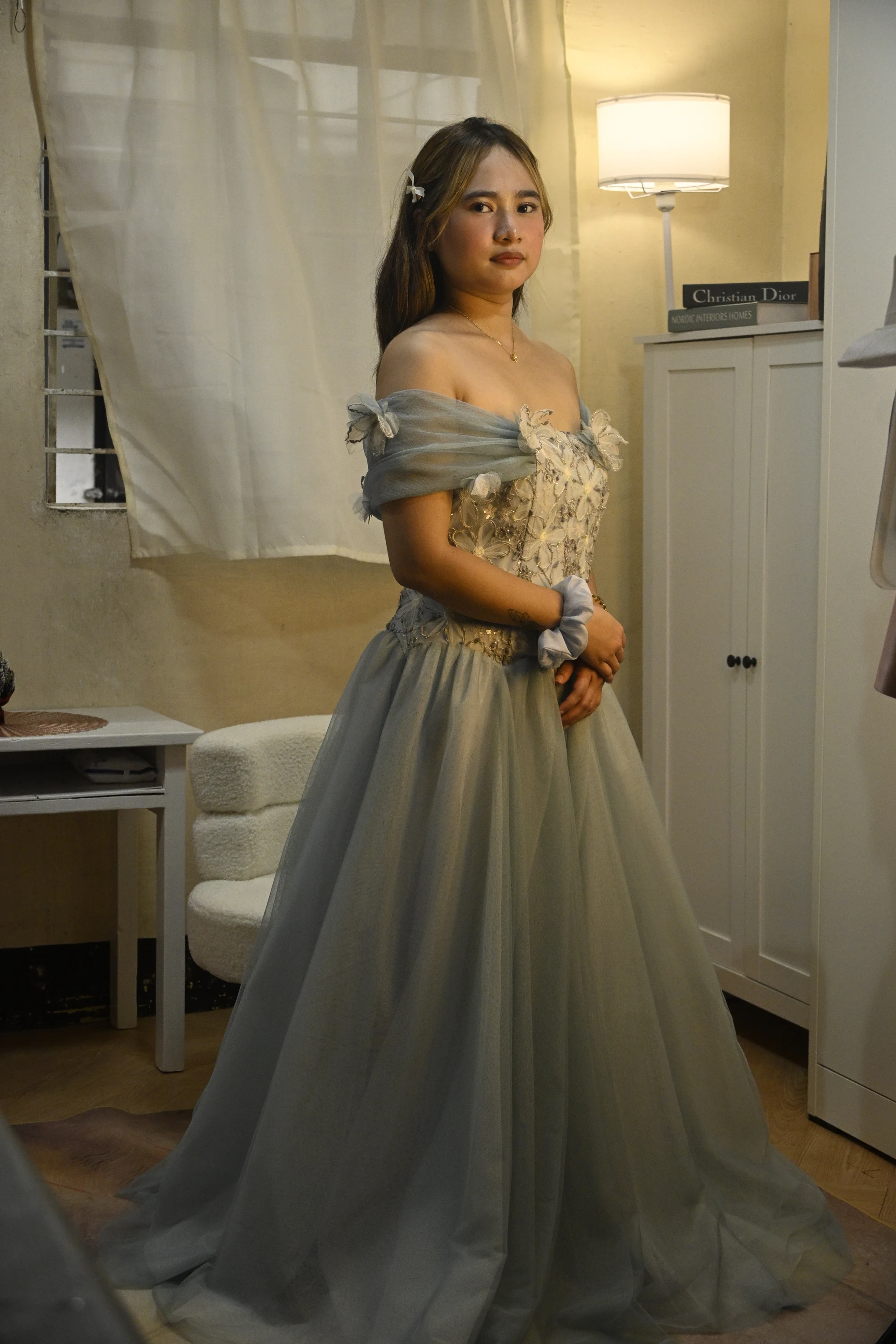 Full-length front portrait of a model in a blue-grey gown with draped off-shoulder sleeves and a floral bodice.