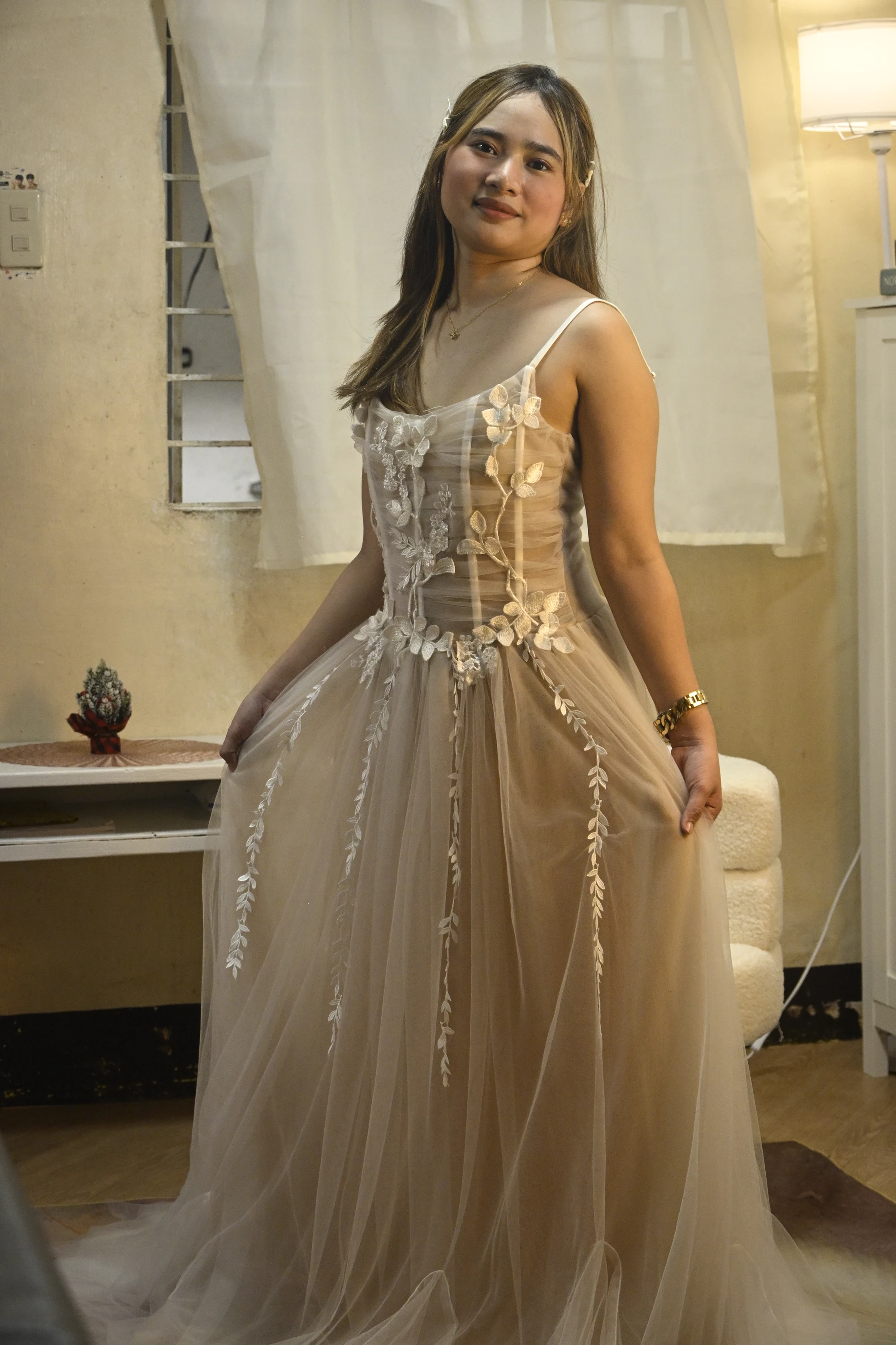 Model holding the skirt of a champagne tulle gown with a sheer corset bodice and trailing floral appliqué.