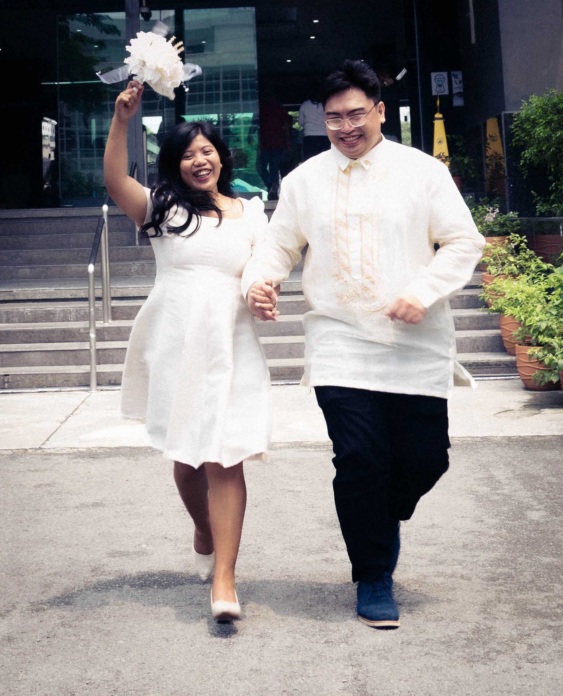 A smiling couple walks hand in hand as the bride raises her white bouquet.