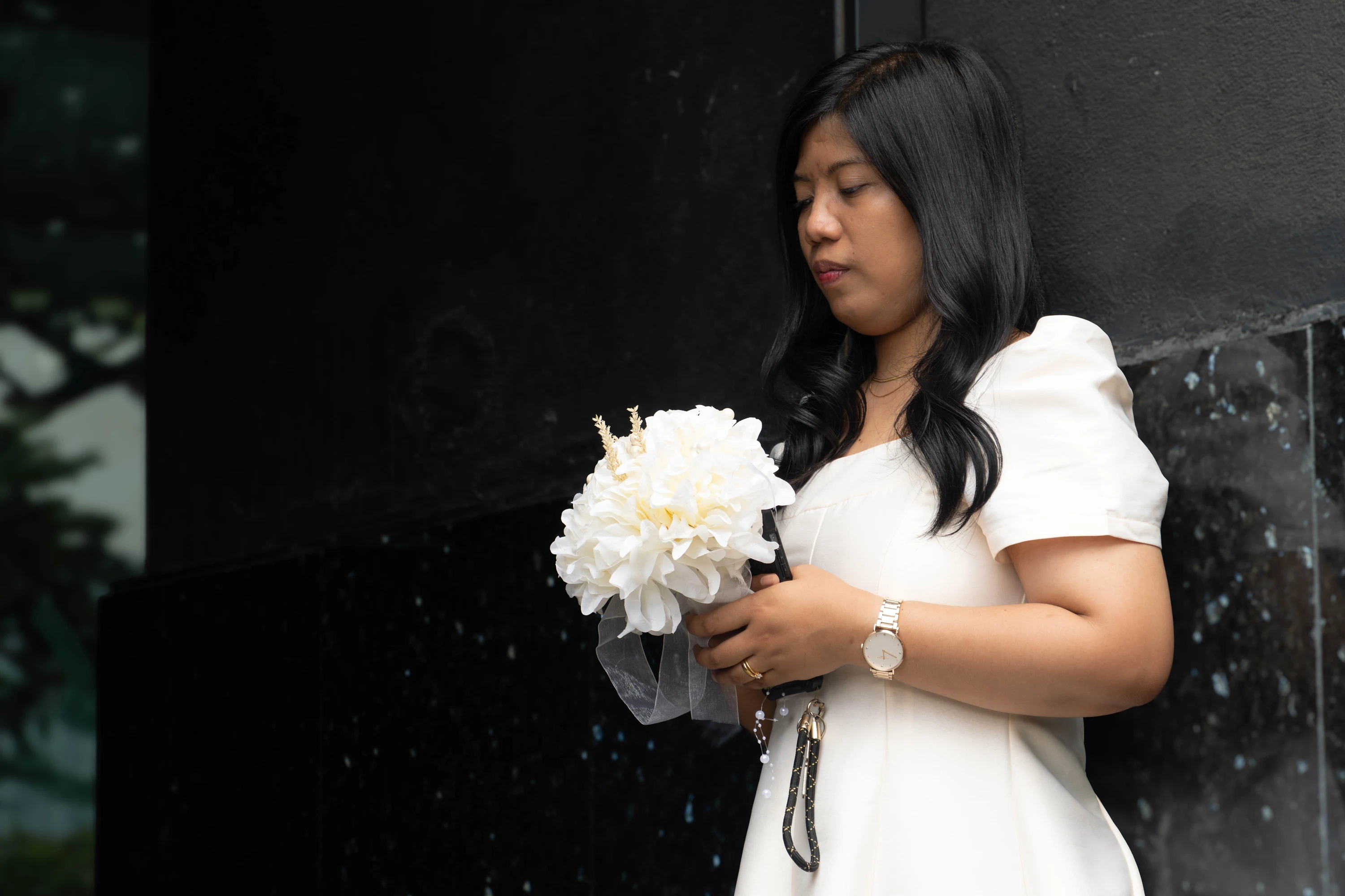 The bride looks toward her white bouquet beside a dark stone wall.