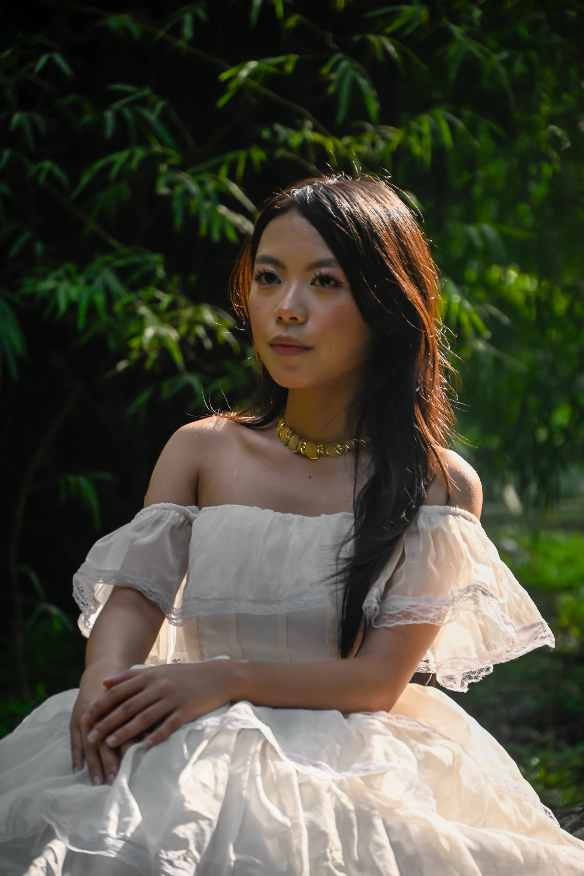Seated portrait in an off-shoulder white gown with hands resting in her lap and sunlight on her hair.
