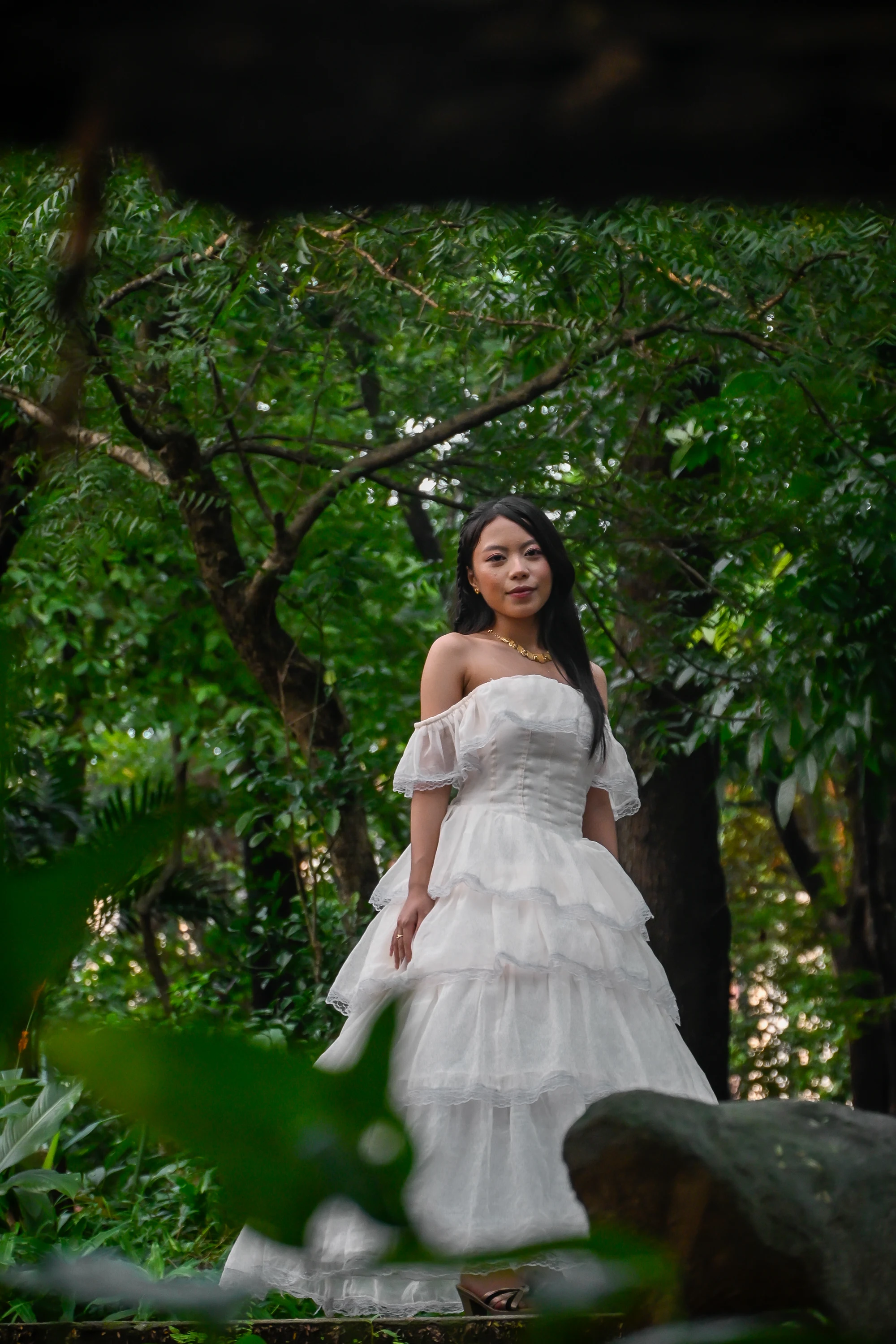 Portrait in a tiered white gown beneath leafy garden branches.
