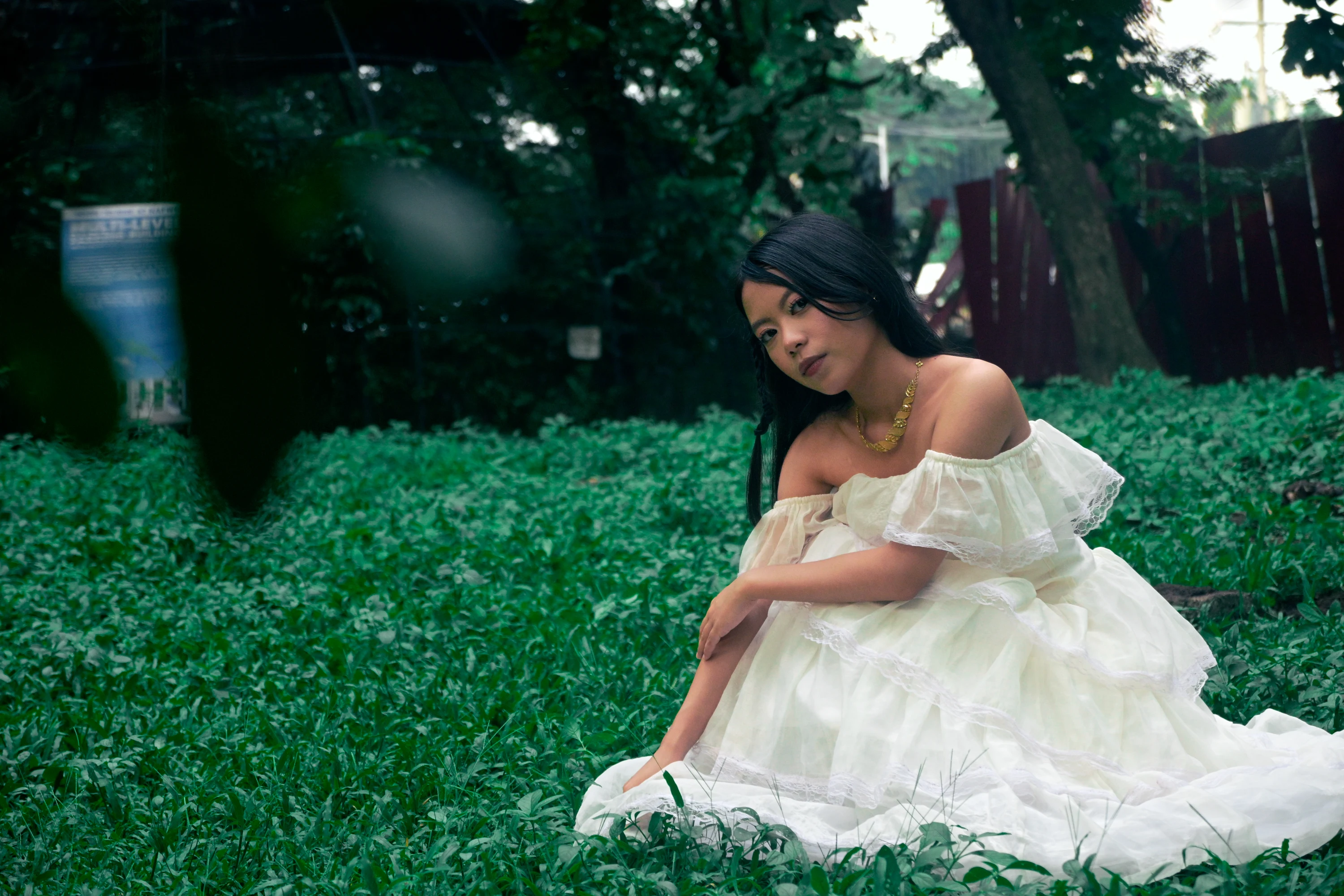 Seated on a green lawn, looking down with a white gown spread beside her.