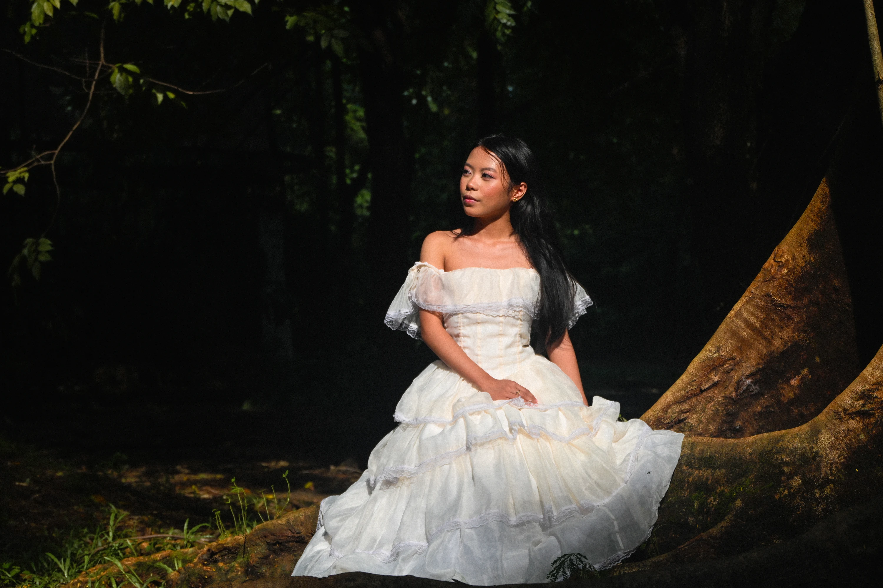 Seated beside a tree in a white gown, lit by a patch of sunlight against a shaded garden.