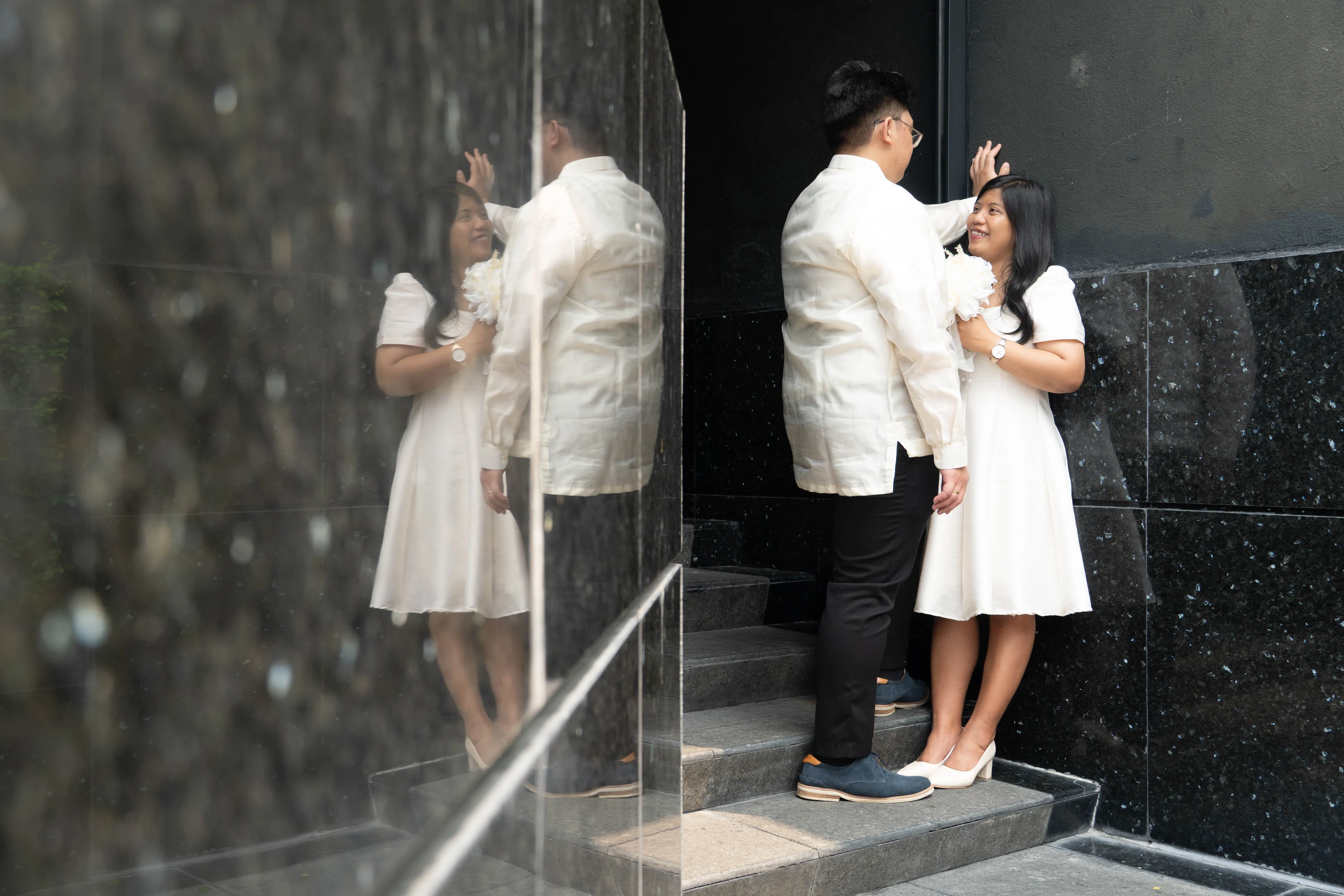 The couple stands on steps with their reflection visible in the adjacent stone wall.