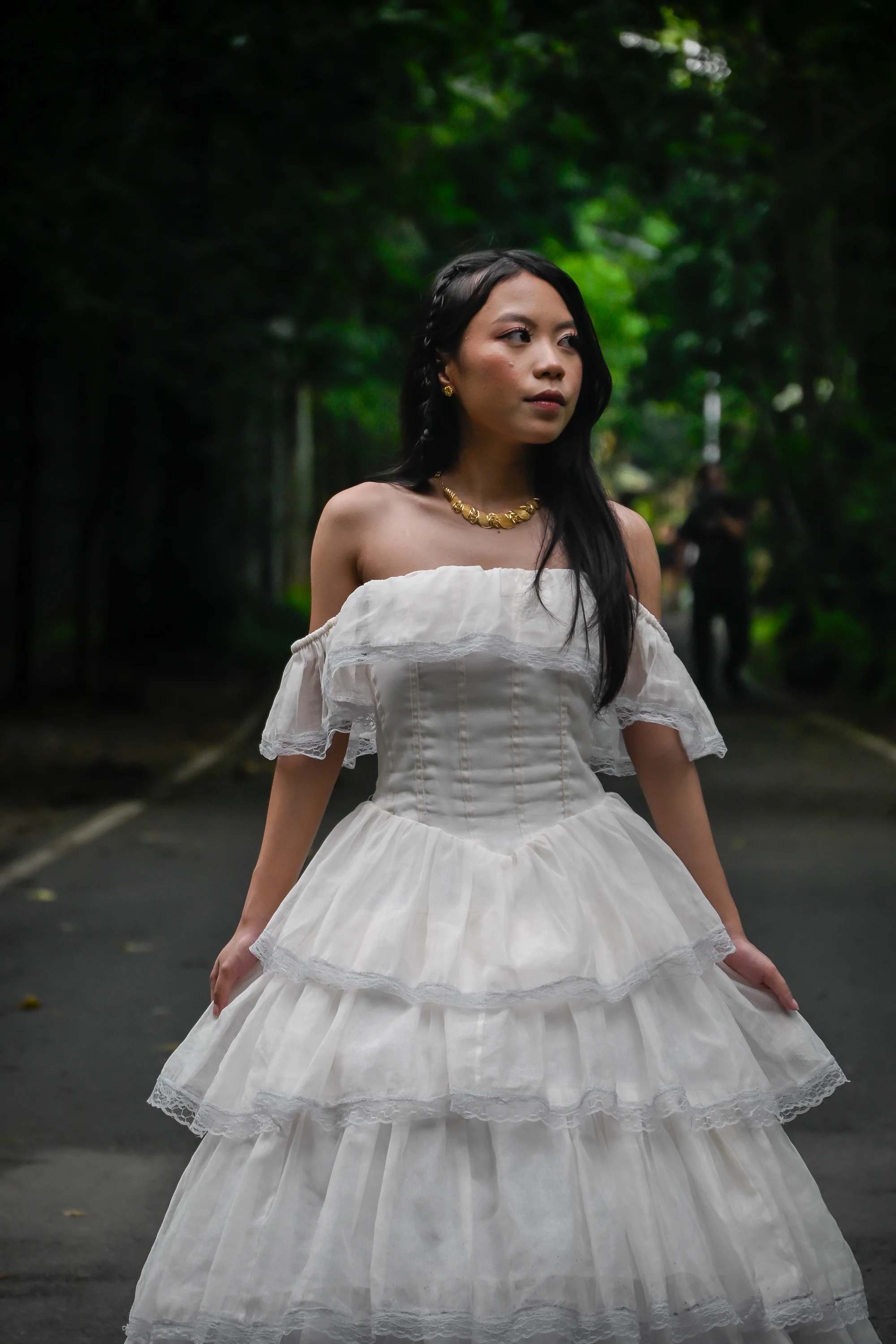 Standing on a garden path in a tiered white gown, holding the skirt and looking to the side.