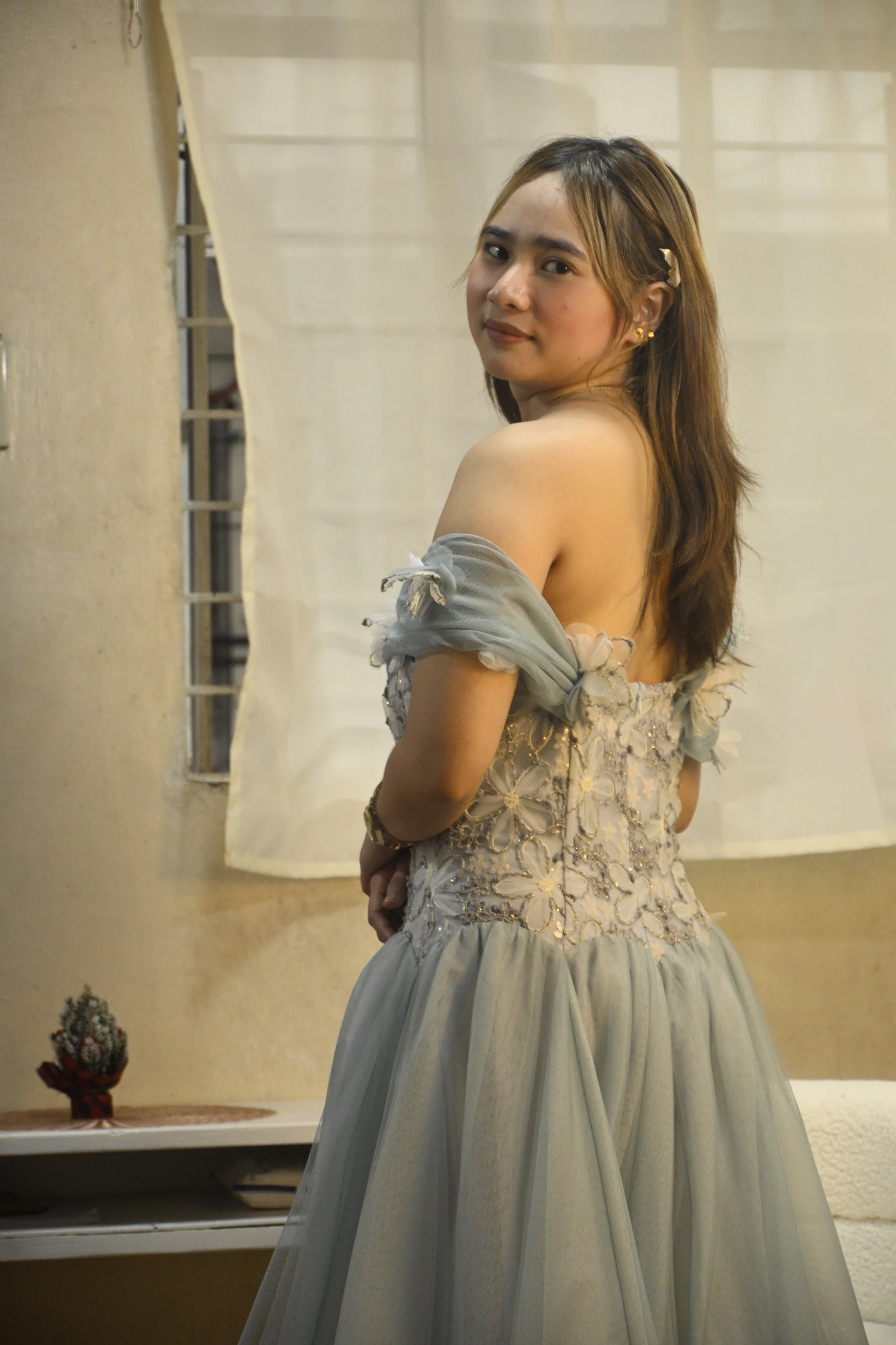Model looking over her shoulder in a blue-grey gown, showing its embroidered back and draped sleeves.