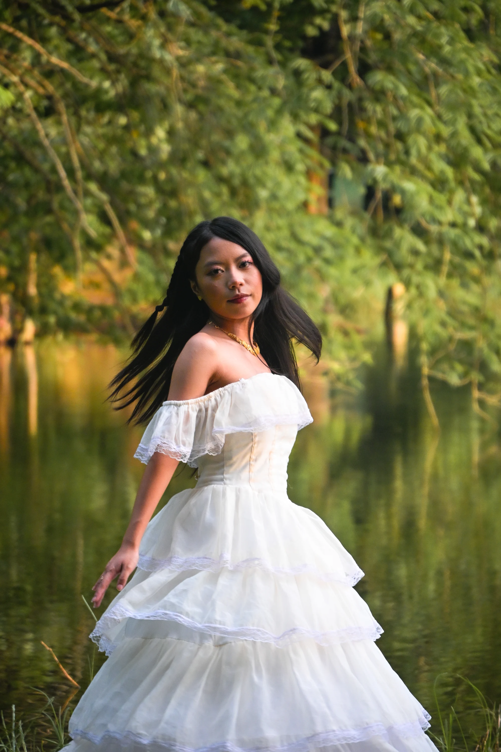 Turning toward the camera in a tiered white gown beside a pond, with her hair sweeping outward.